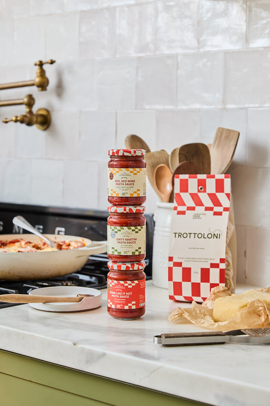 Three jars of Saturday Night Pasta sauce stacked vertically next to a red and white checkered bag of the same brand on a kitchen counter, with a pan of pasta and kitchen utensils in the background.