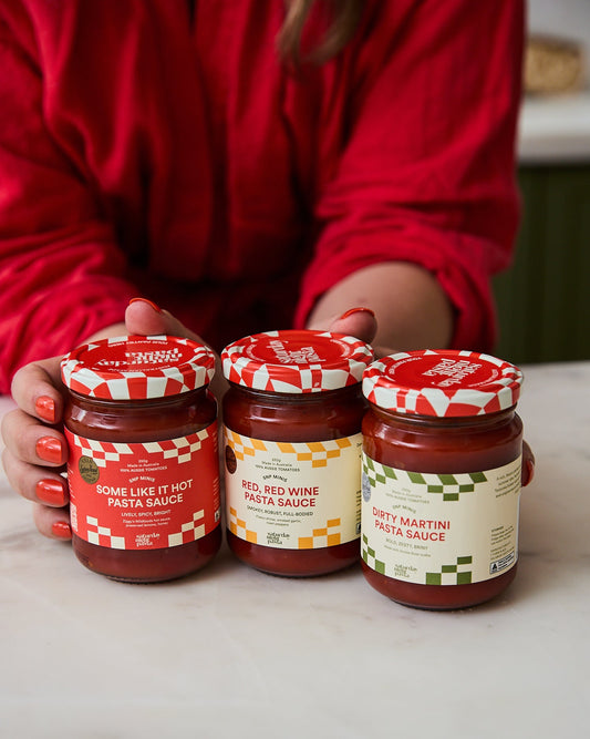 Three glass jars of pasta sauce are held by a person wearing a red top on a light marble countertop. The labels read 'Some Like It Hot Pasta Sauce', 'Red Wine Pasta Sauce', and 'Dirty Martini Pasta Sauce'.