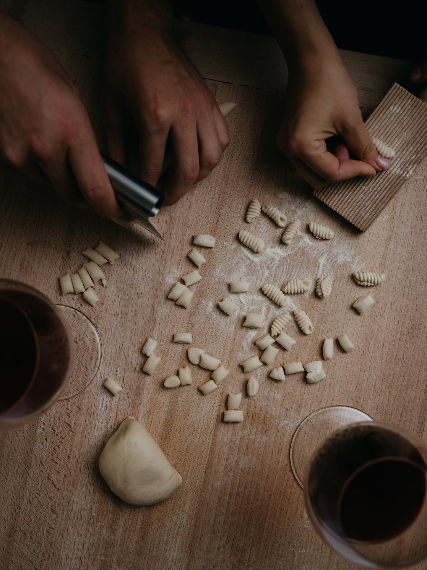 Hands making pasta on a wooden surface with glasses of red wine.