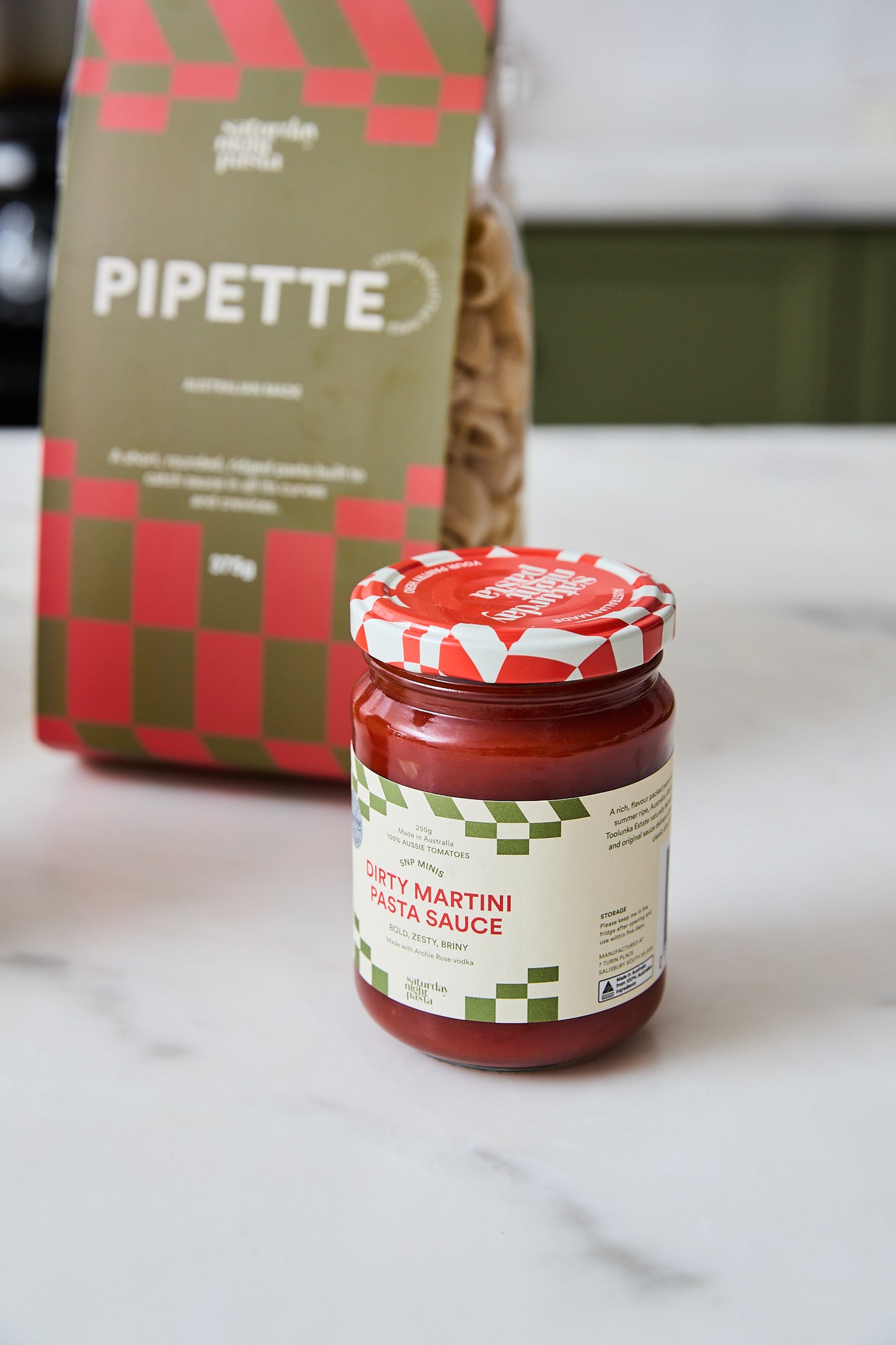 A jar of Dirty Martini Pasta Sauce in the foreground on a white marble countertop, with Pipette pasta box standing behind it.