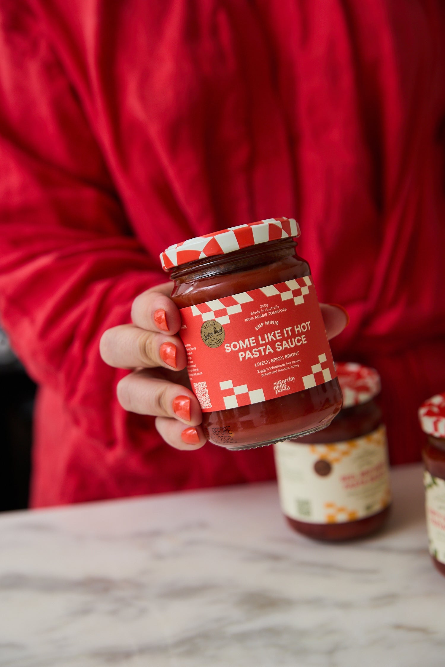 A hand holds a glass jar of pasta sauce labeled 'Some Like It Hot Pasta Sauce' against a red background, with other jars on a marble surface.