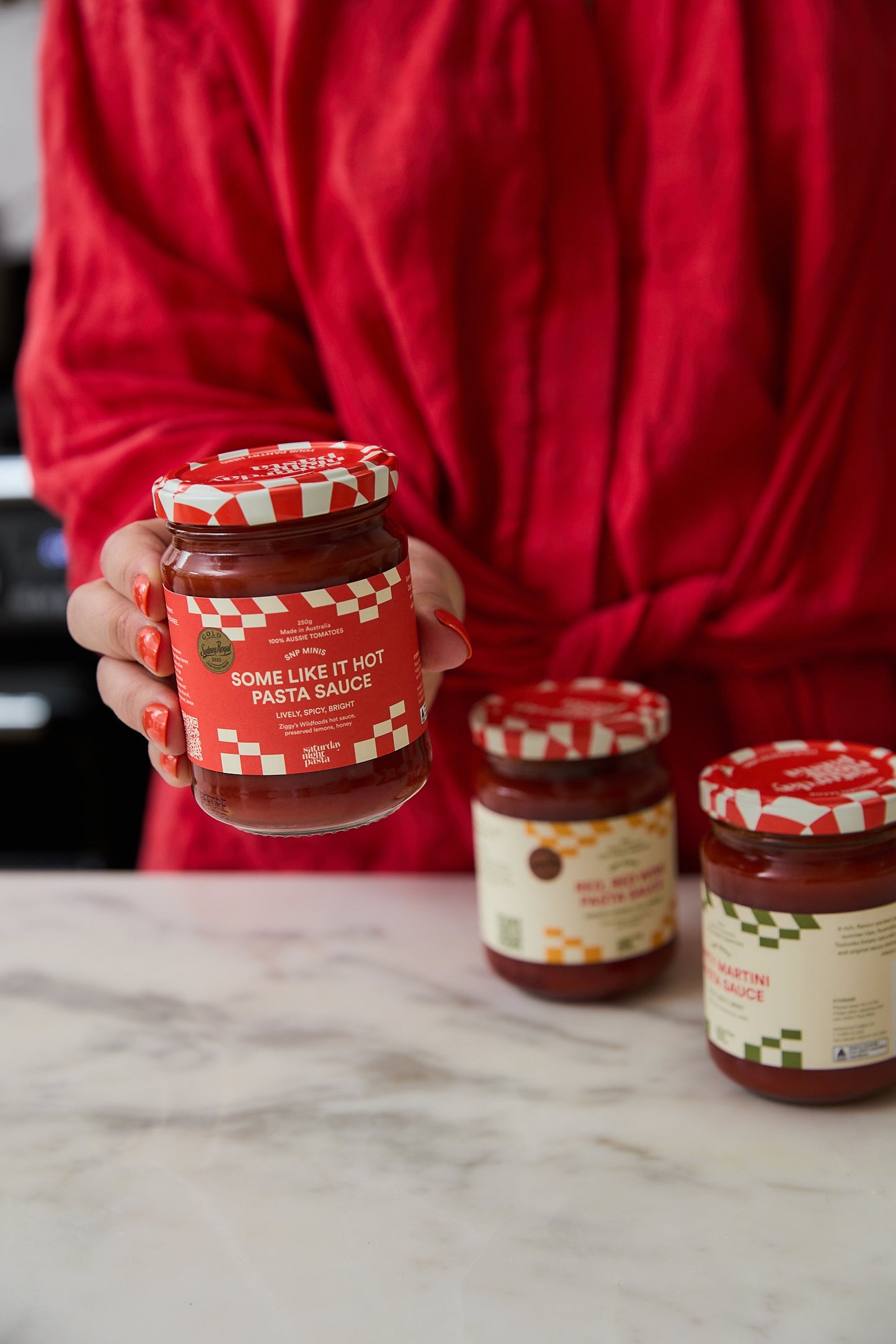 A person in a red top holds a glass jar of pasta sauce in front of two other jars on a marble countertop.