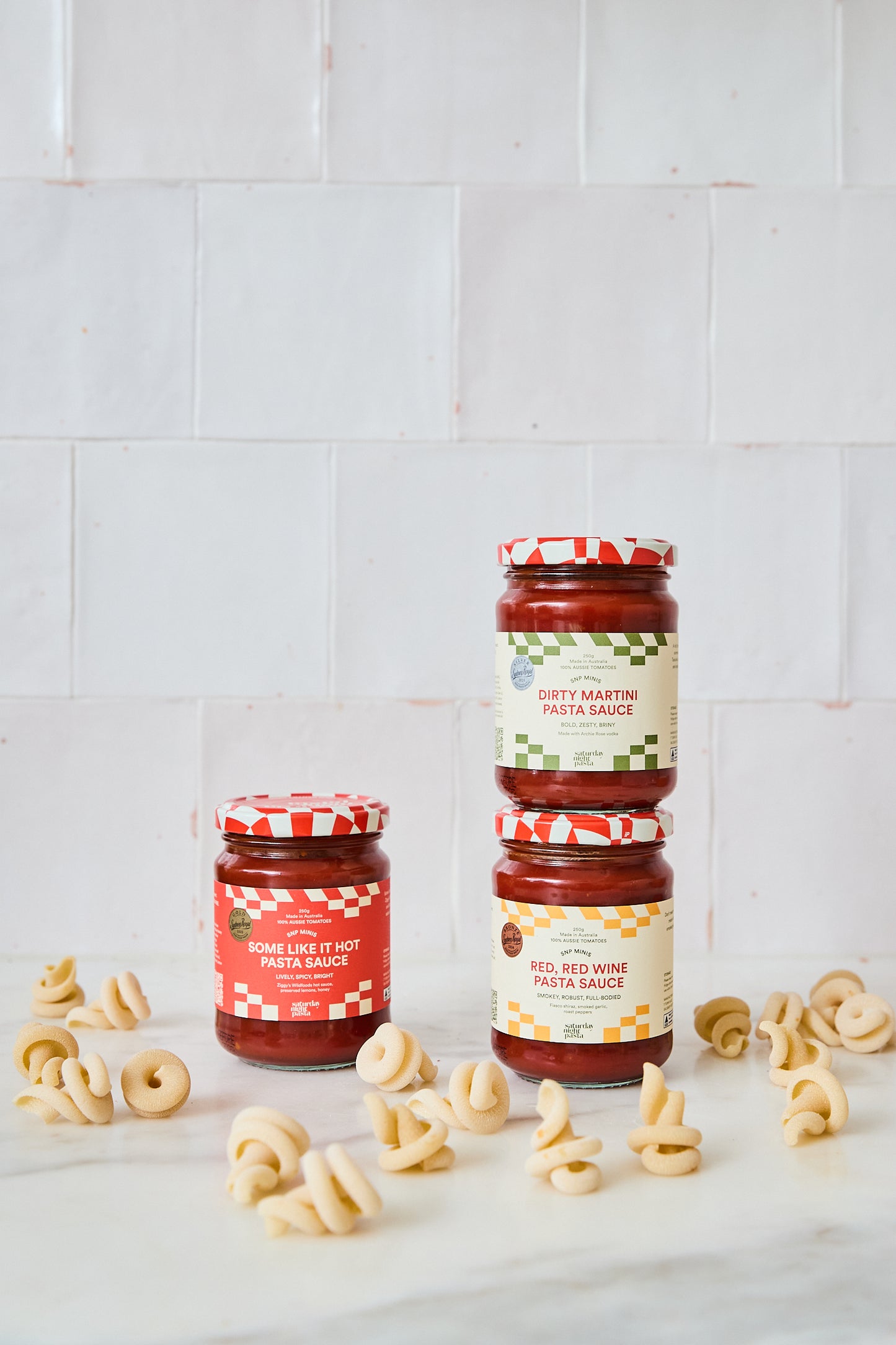 Three jars of pasta sauce arranged on a white marble surface with loose penne pasta scattered around, against a white tiled background.