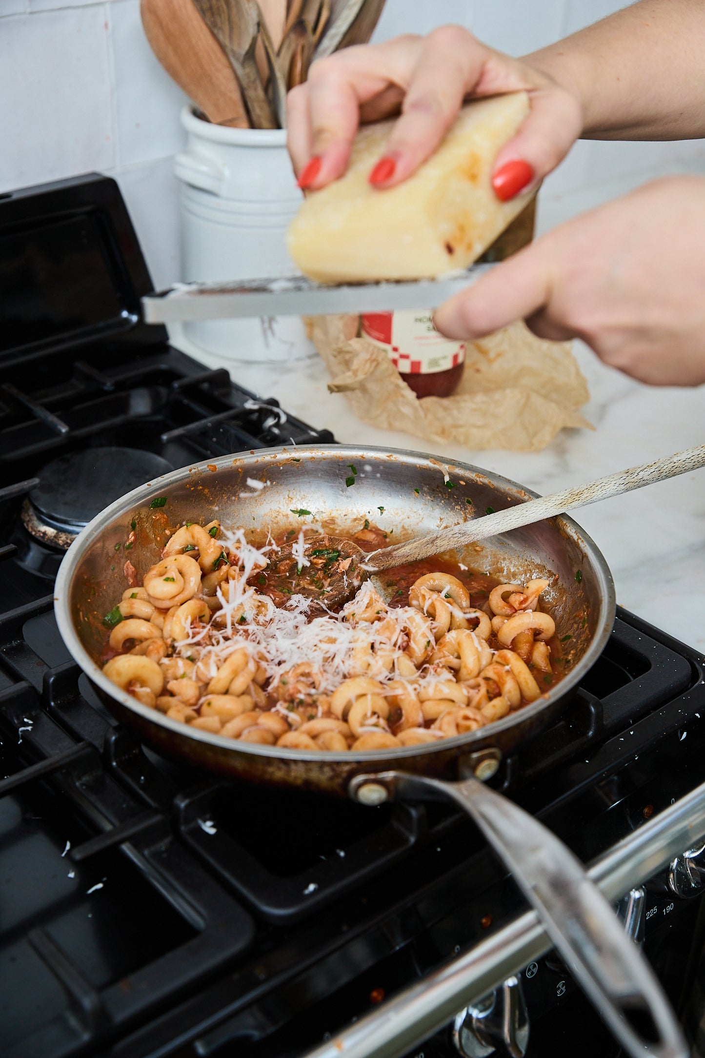 A stainless steel pan filled with pasta on a black gas stovetop, with a person's hands grating a block of cheese over the dish.