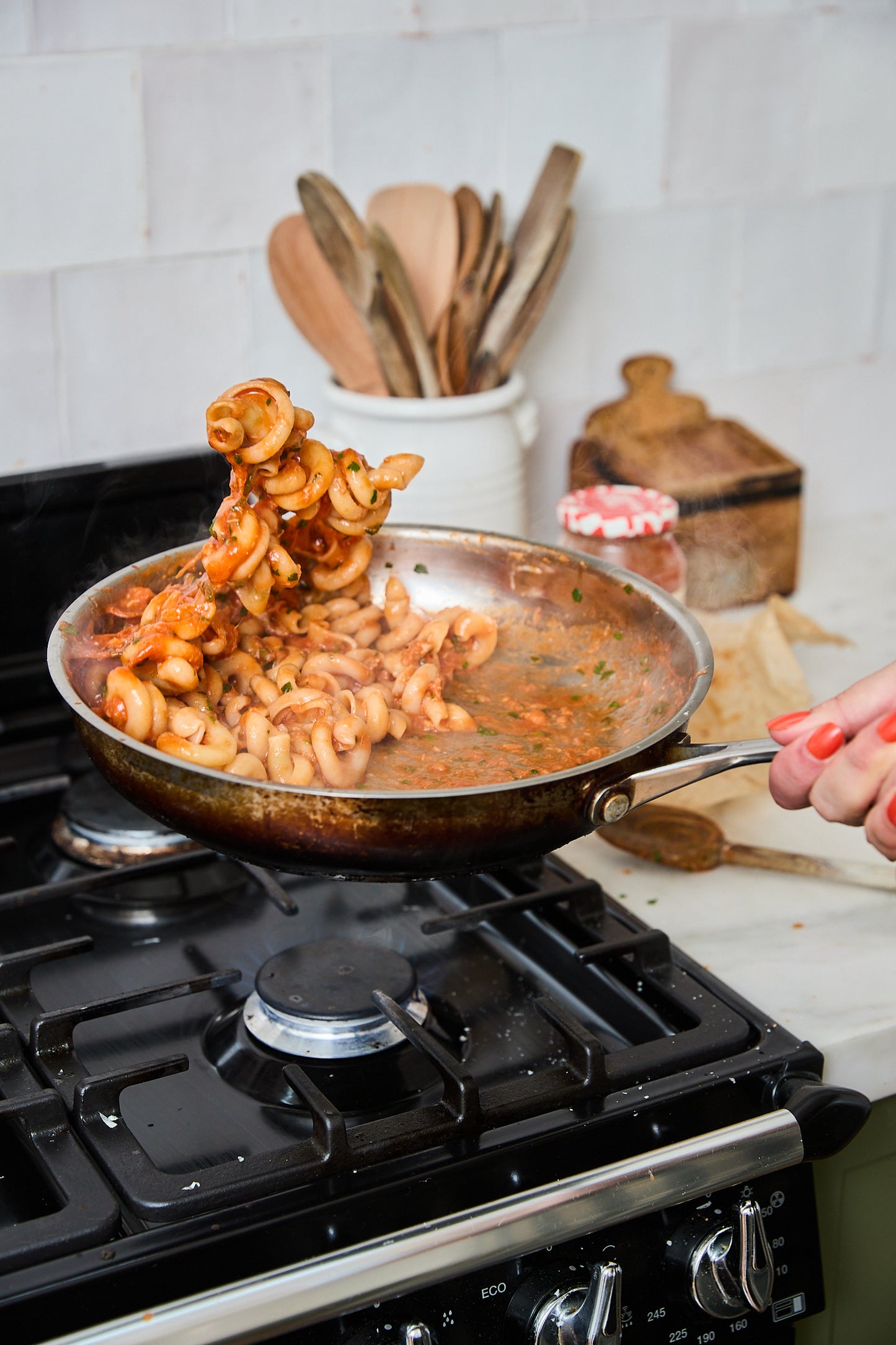 A person tosses cooked pasta in a stainless steel frying pan over a black gas stove in a kitchen setting.