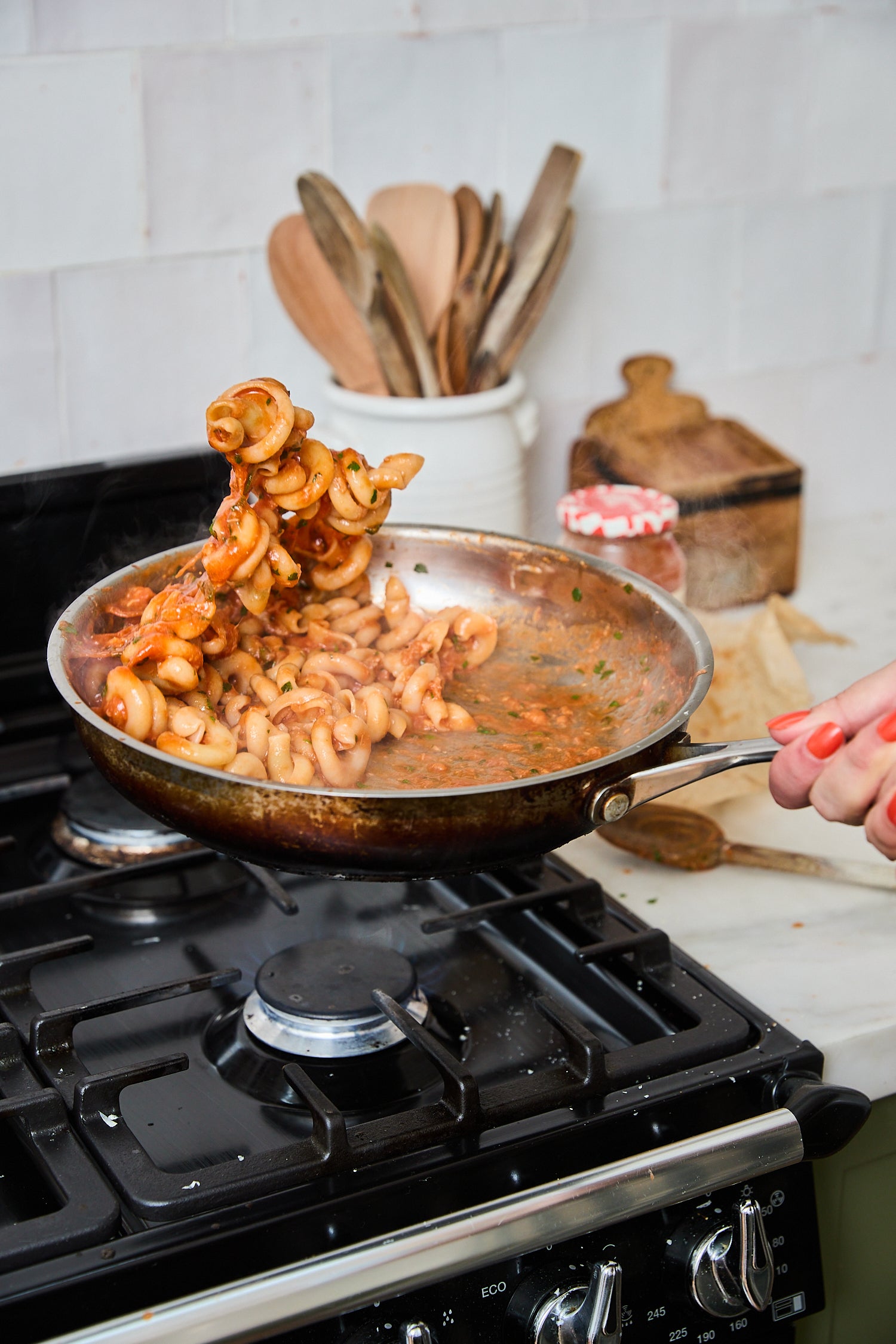 A person tosses cooked pasta in a stainless steel frying pan over a black gas stove in a kitchen setting.