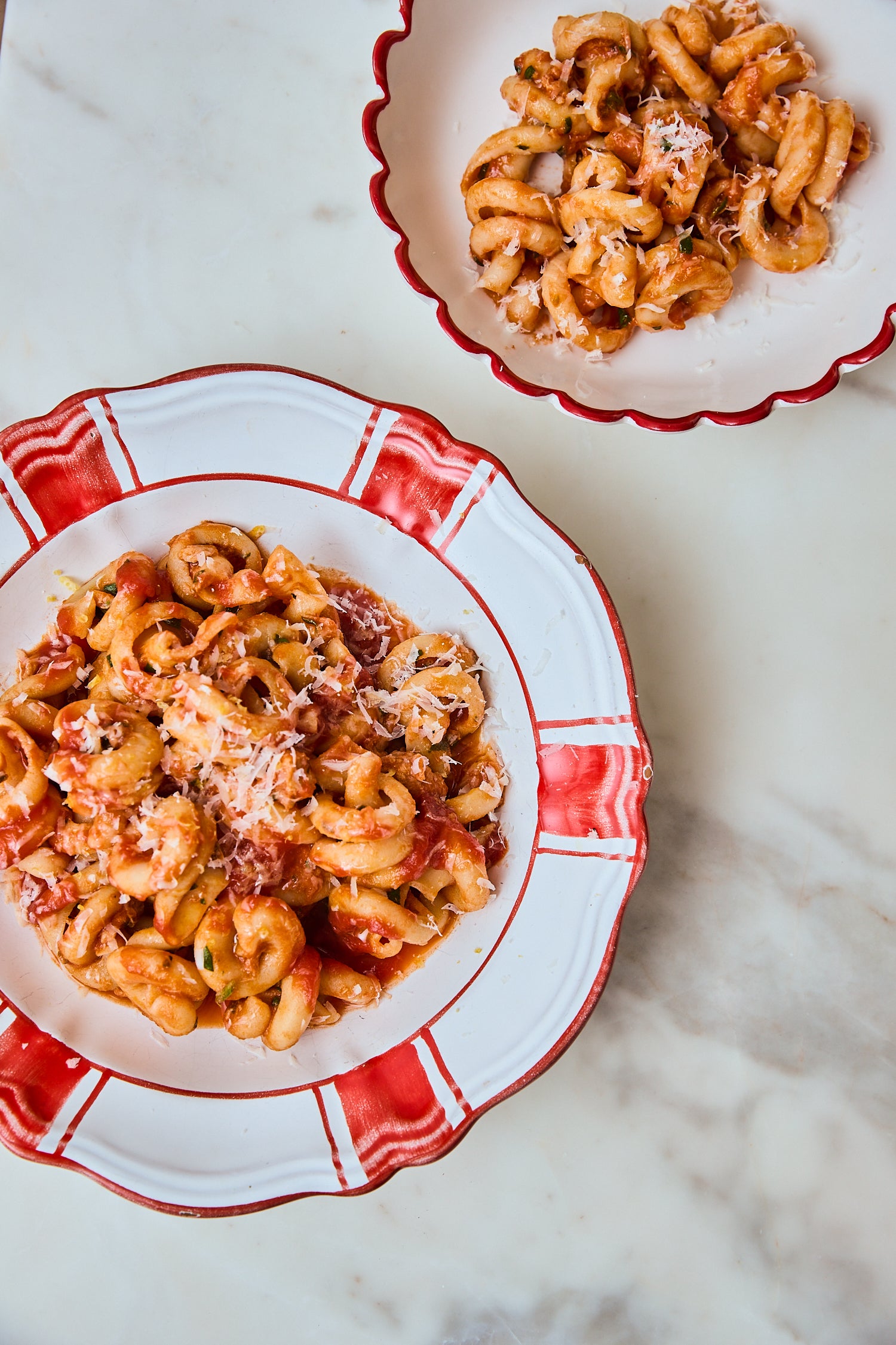Two scalloped white ceramic bowls with red trim, filled with pasta, set on a light marble surface.