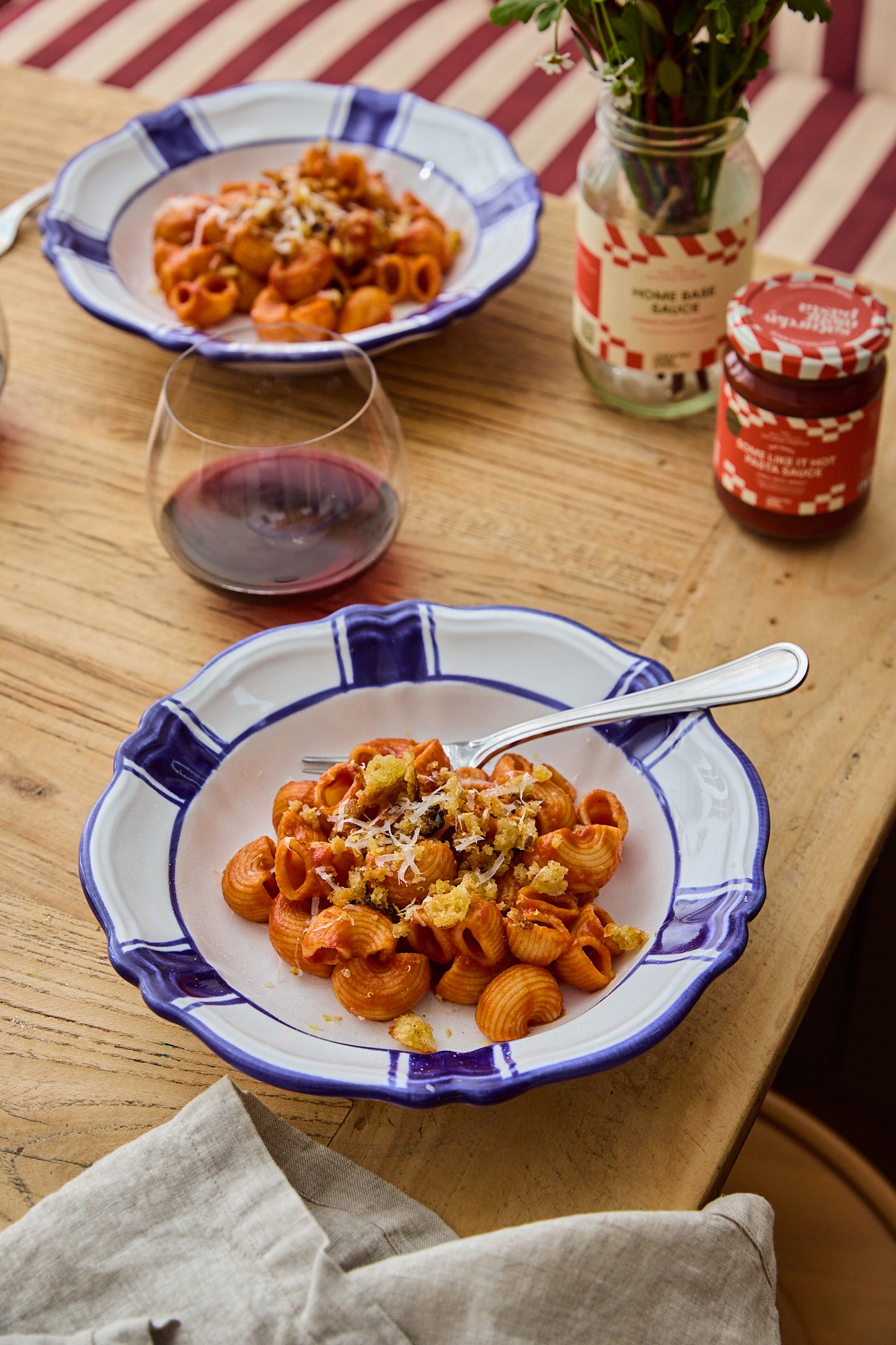 A bowl of pasta with a blue and white patterned plate on a wooden table, with a second bowl, a glass of red wine, and jars in the background.