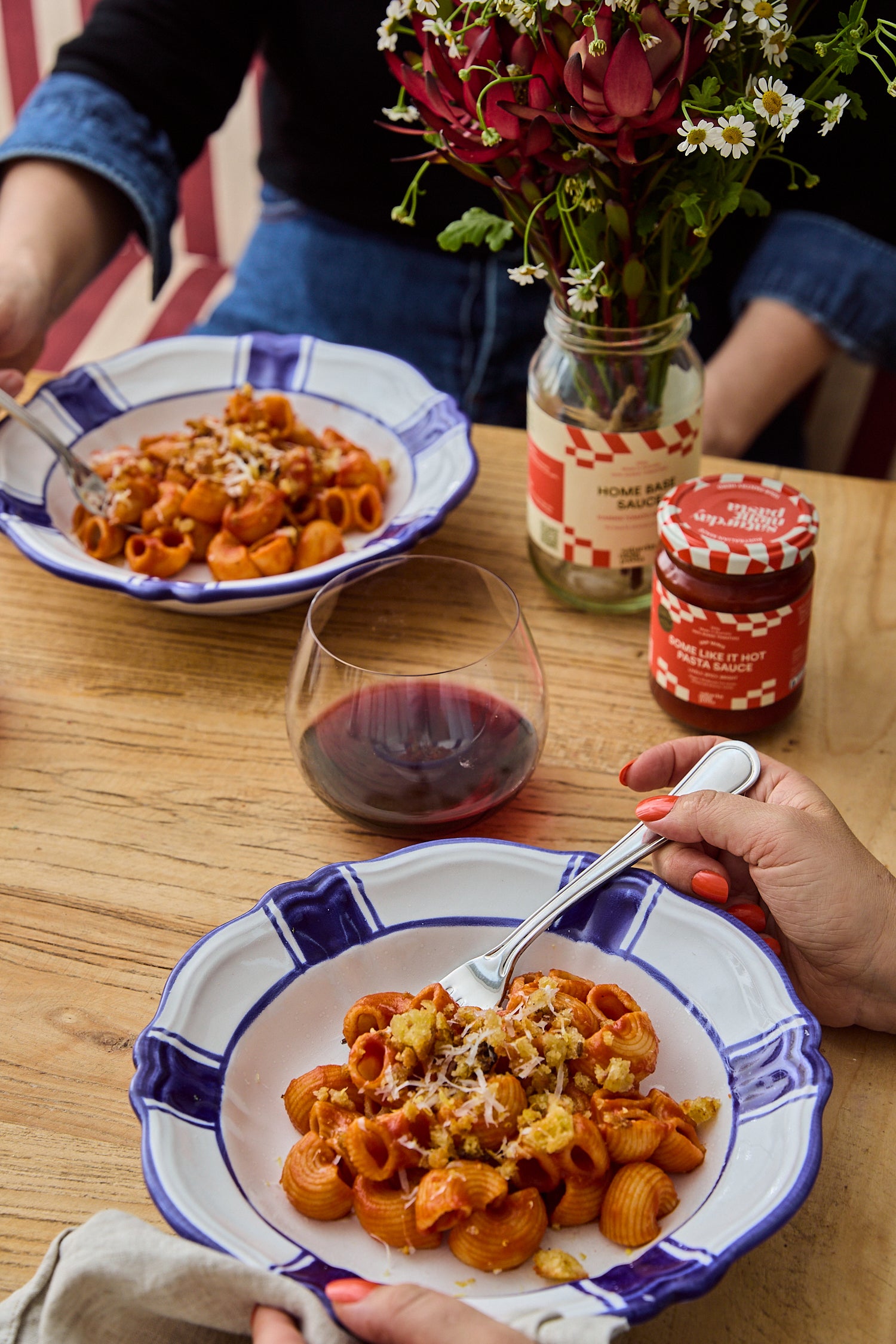 Two people seated at a wooden table sharing a bowl of pasta with red sauce, cheese, and garlic. A glass of red wine, two jars, and a vase of flowers are on the table.