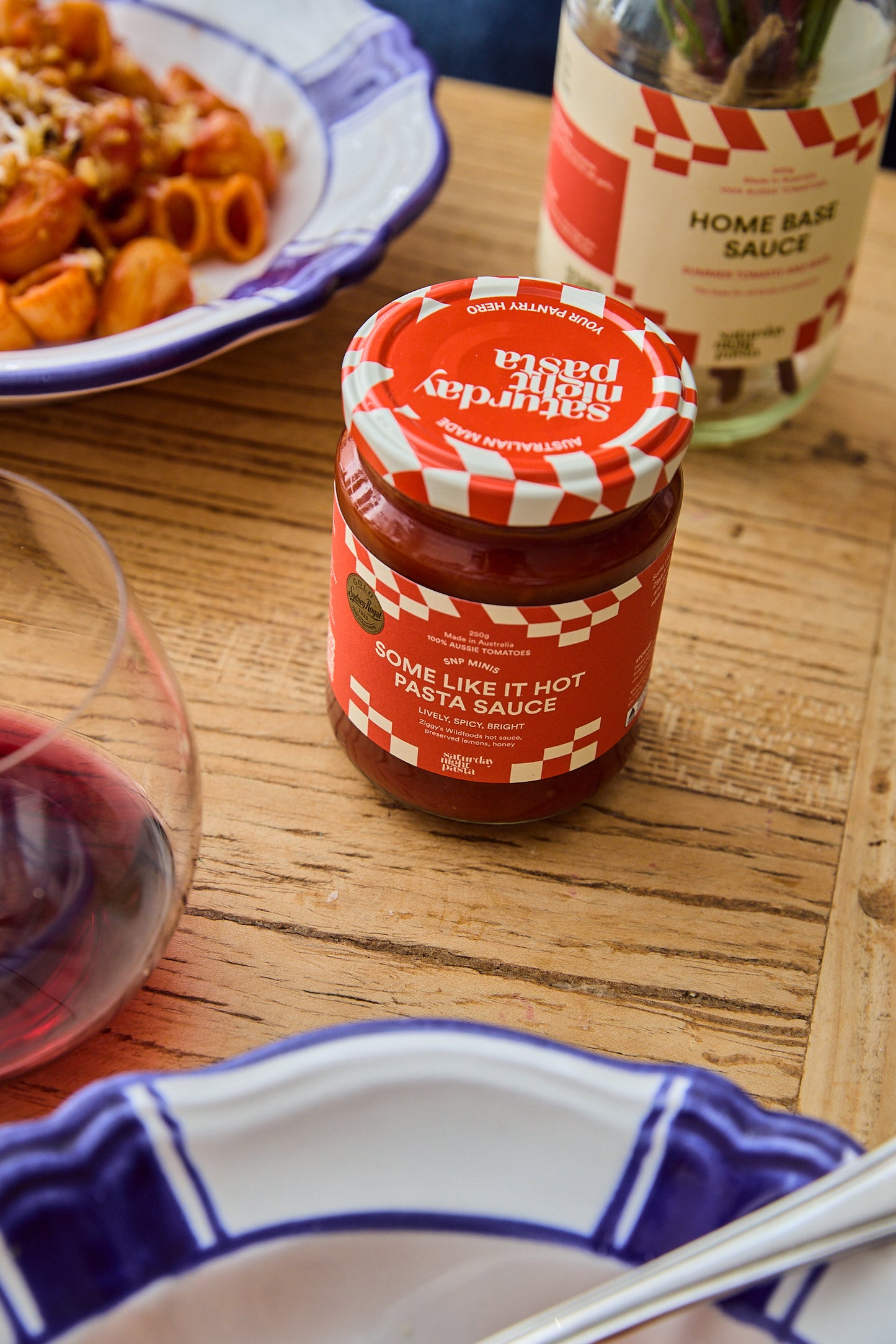 A jar of Saturday Night Pasta Sauce sits on a wooden dining table, surrounded by plates of food and a glass of red wine. The label reads 'SOME LIKE IT HOT PASTA SAUCE' and the brand 'Saturday Night Pasta Sauce' is visible on the lid.