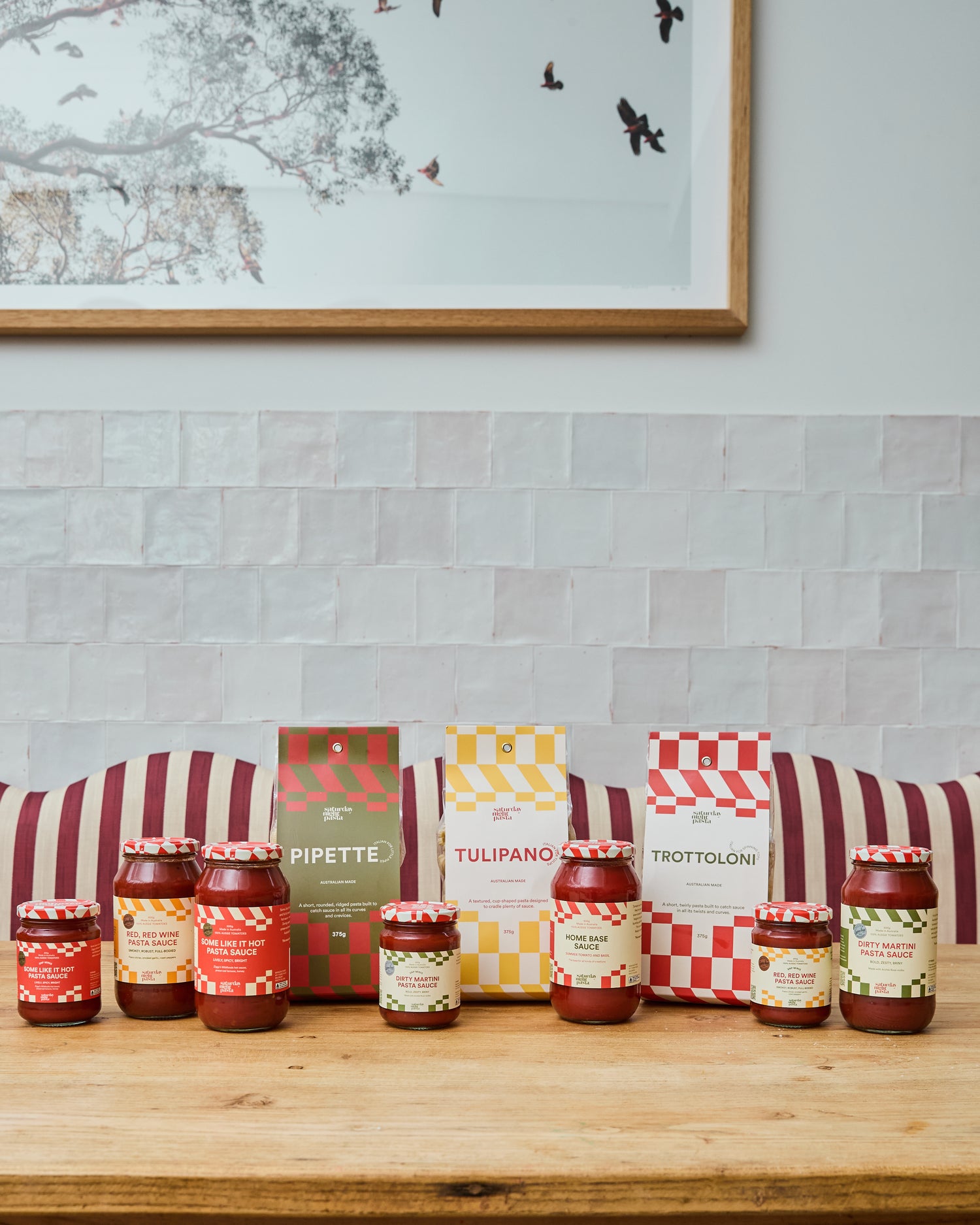 A collection of gourmet pasta and sauce products from the brand Saturday Night Pasta, arranged on a wooden table in front of a white brick wall and a framed print. The products include jars of red sauce and boxes of pasta in red, green, and yellow checkered packaging.