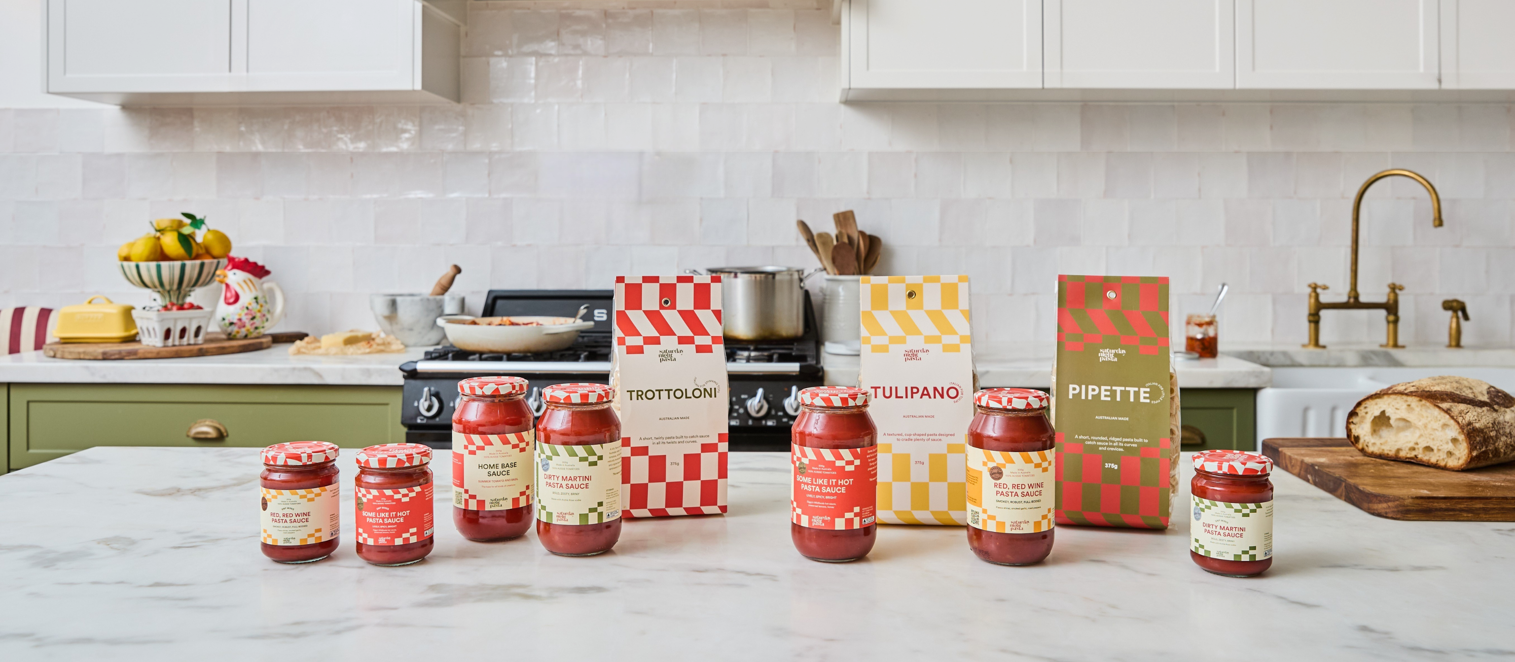 A collection of pasta sauce jars and slow-dried pasta from the brand Saturday Night Pasta, arranged on a white marble kitchen counter with a white tiled backsplash and green cabinets in the background.